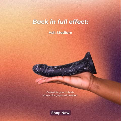 Back in full effect: Ash Medium. Crafted for your body. Curved for g-spot stimulation. A dark-skinned hand holds a black and grey curved dildo. 
